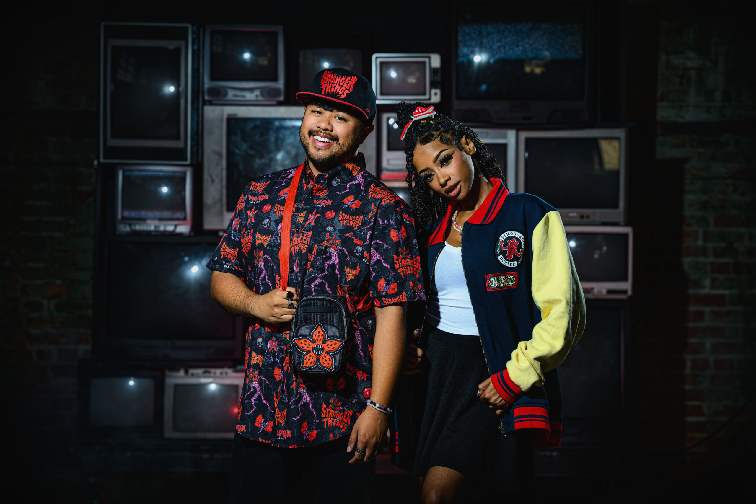 male and female modeling Stranger Things merchandise including printed button-up, varsity jacket, bag, trucker hat and scrunche.