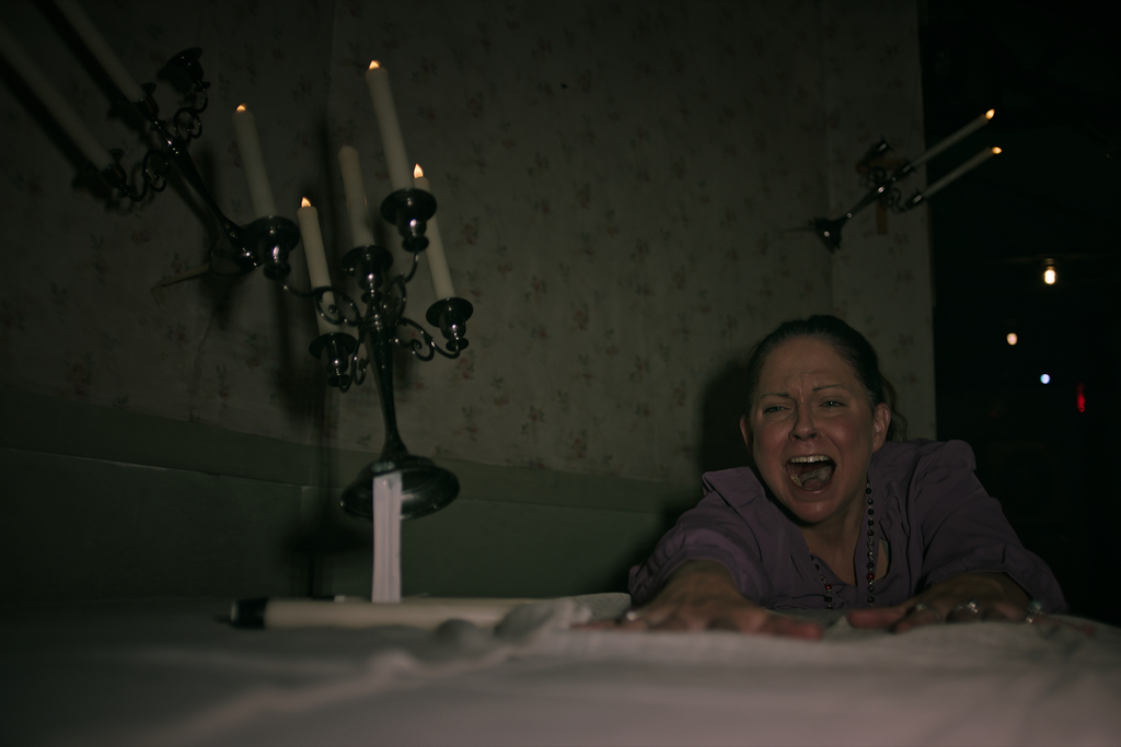 Women in dimly lit room, screaming and reaching across table. Candelabras float around her.