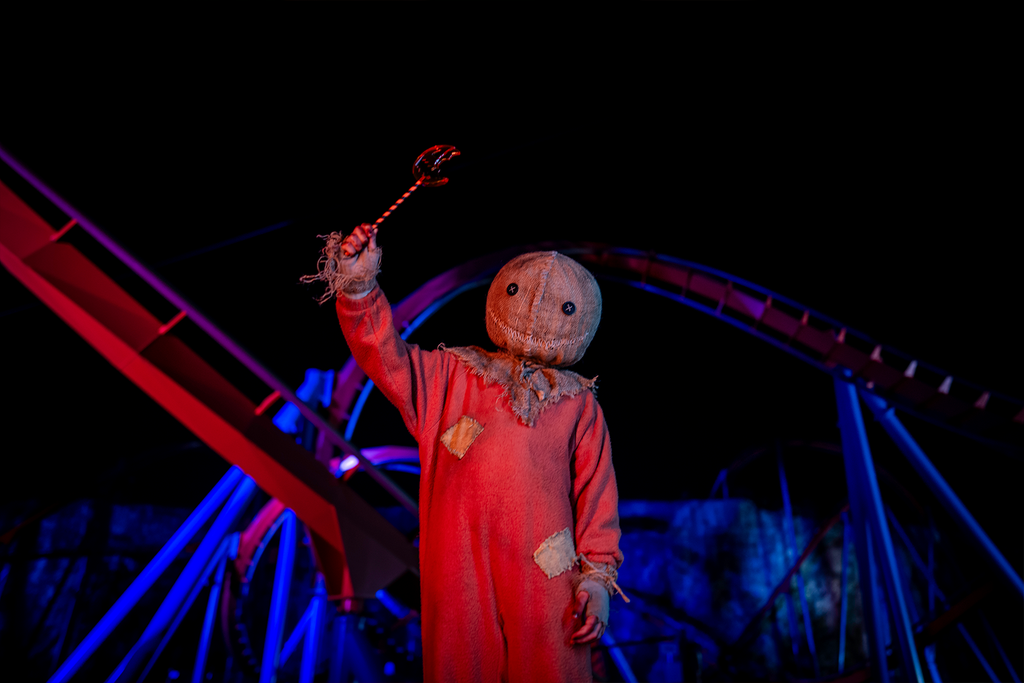 Sam character from Trick 'r Treat (orange jumpsuit and burlap sack mask) holds up sharp lollipop in front of roller coaster